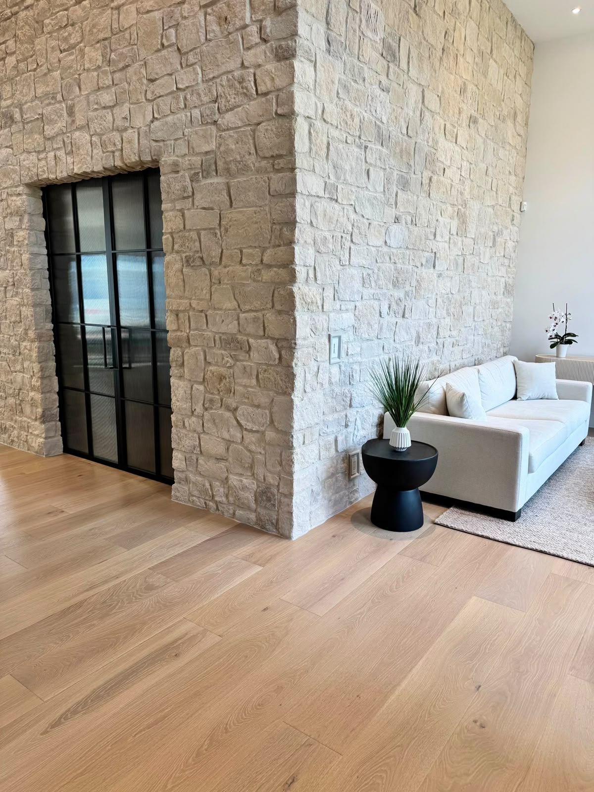 A stone wall wrapping a corner beside black steel and glass doors, with a white sofa and a black side table to the right.