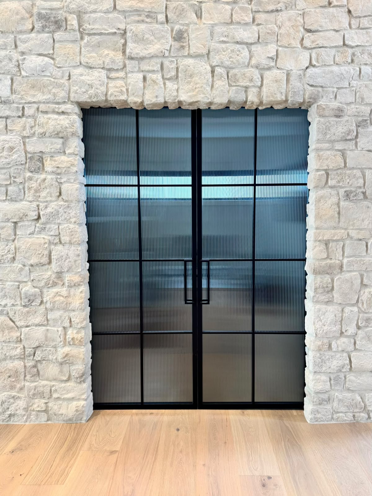 A pair of black steel doors with ribbed glass set into an opening in a stone wall, over an oak floor.