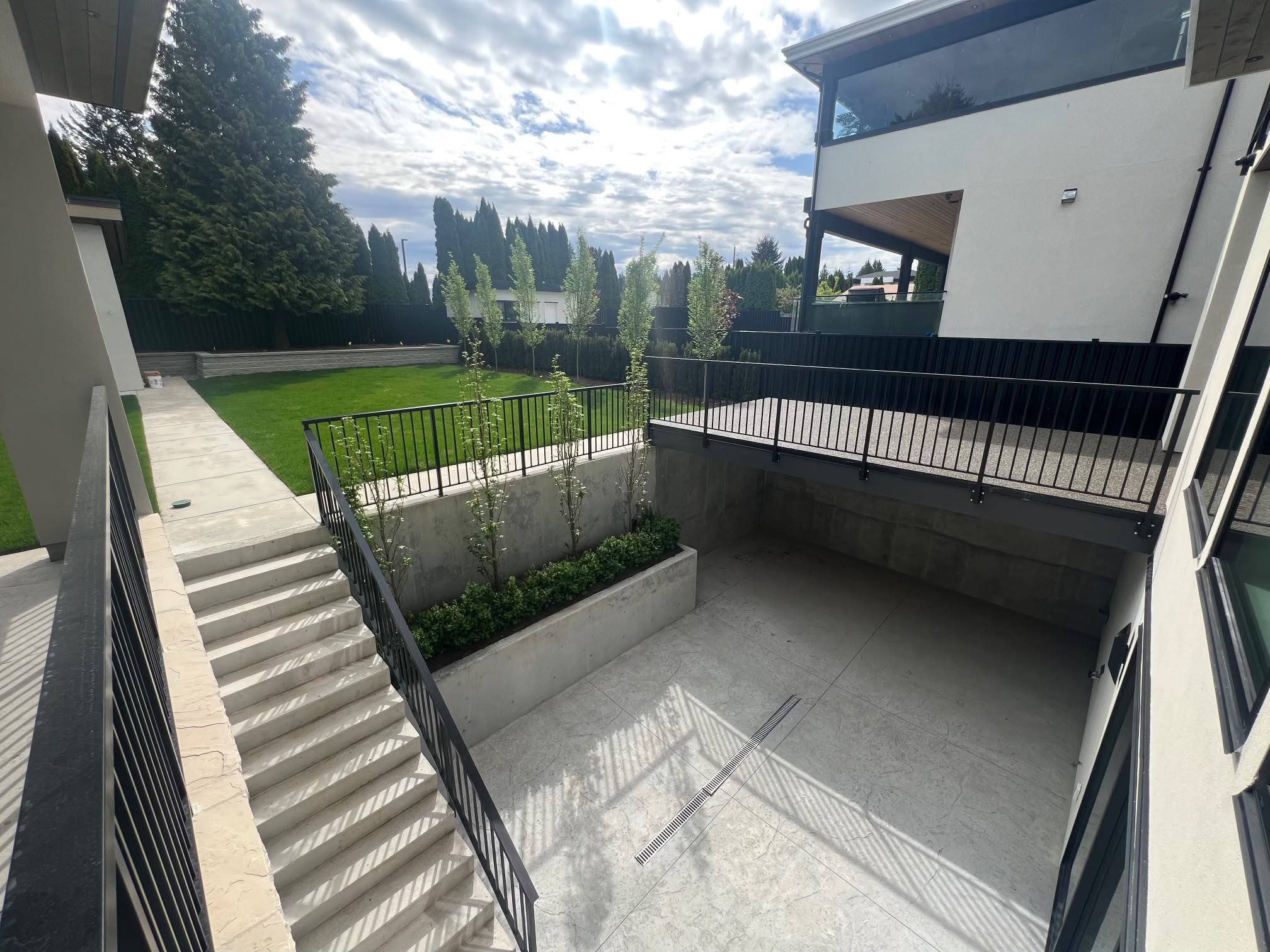 The back of the house: a concrete stair beside a planted wall down to a courtyard, the lawn beyond and a deck with a black rail above.