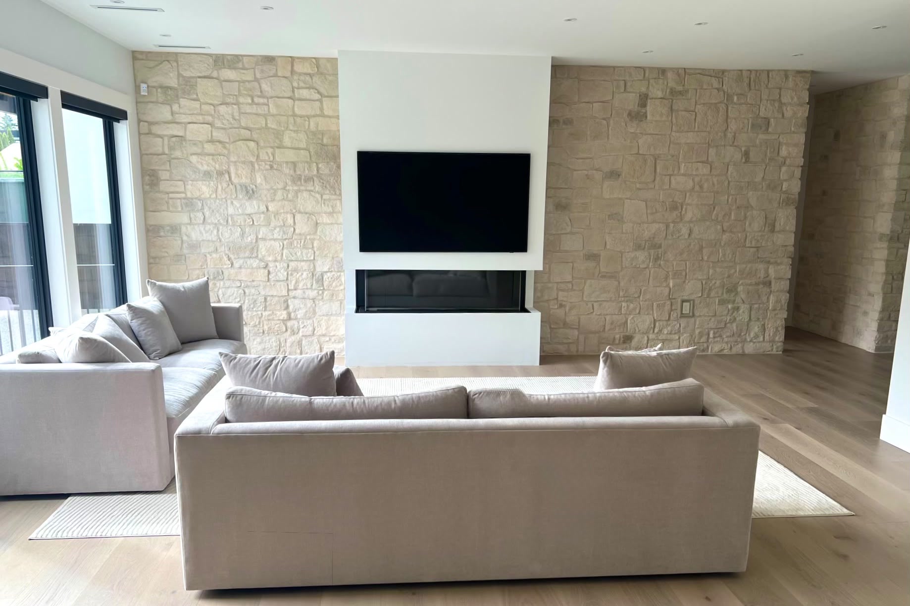 The living room: stone walls either side of a white panel holding a television above a linear fireplace, with two grey sofas.