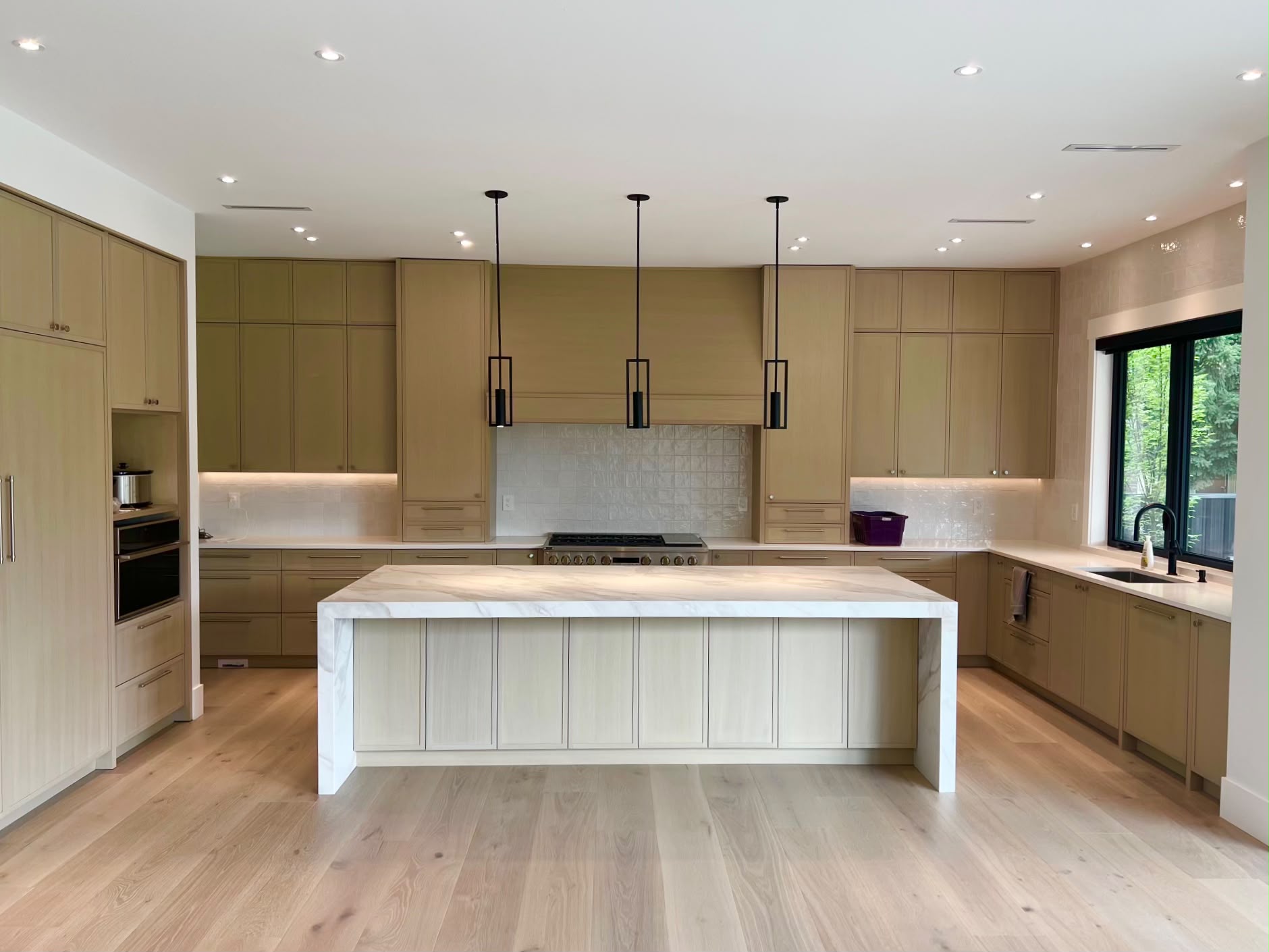 The kitchen: light wood cabinets to the ceiling, a tiled wall behind the range and a waterfall island with a veined top under three black pendants.