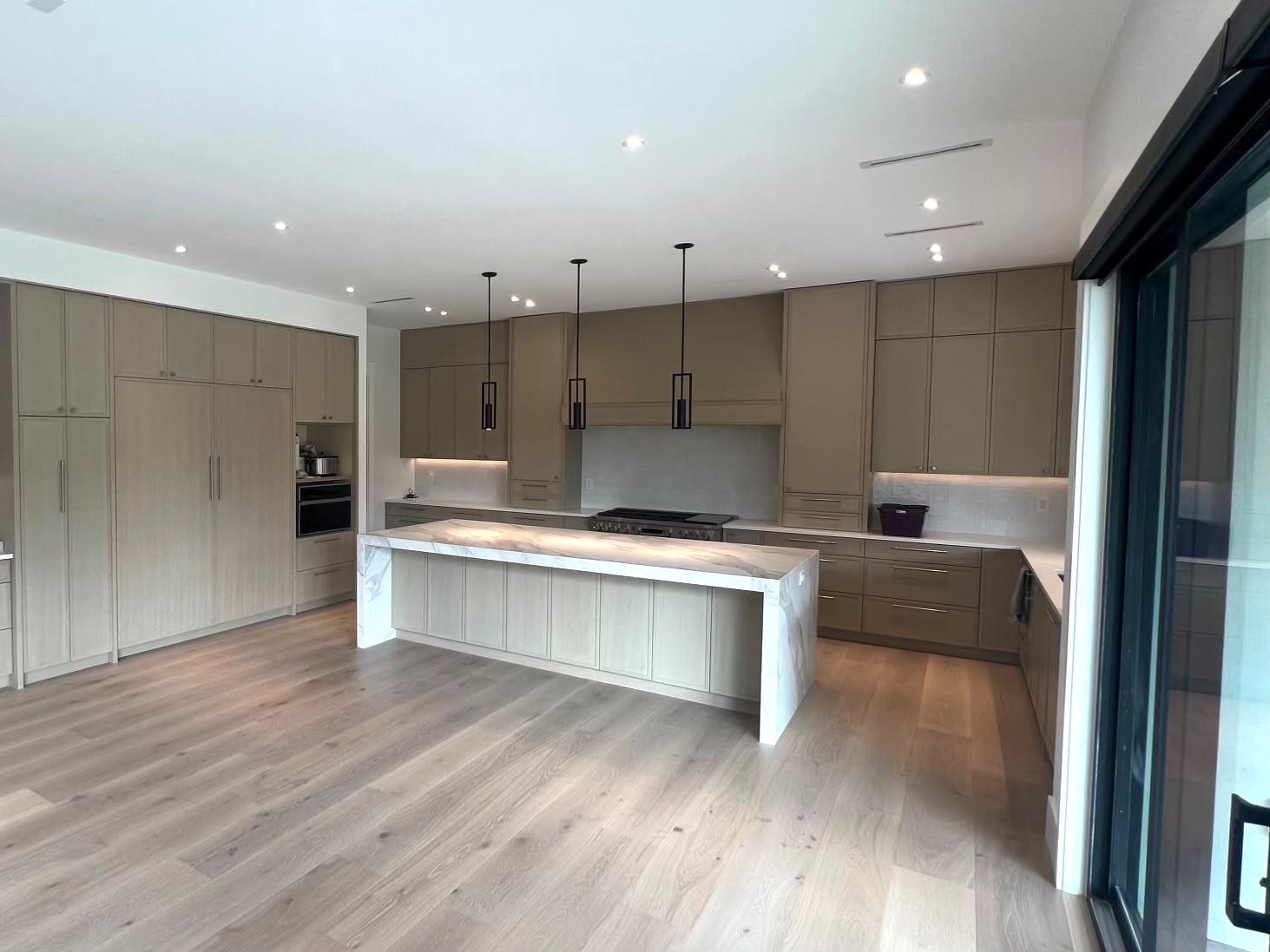 The kitchen from the corner: light wood cabinets, a range under a hood, a waterfall island and a sliding glass door at the right.